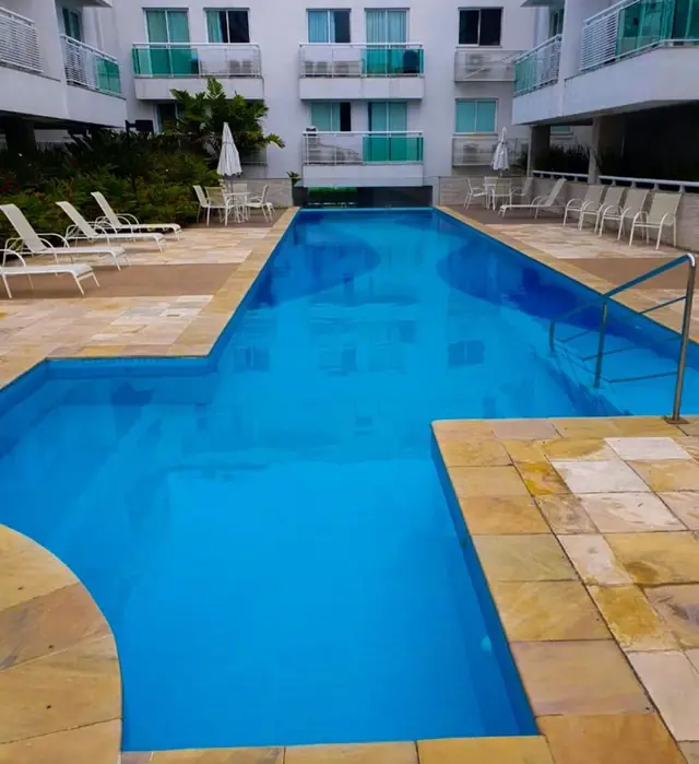 Piscina de condomínio com a água azul e cristalina, cercada de pedra clara e espreguiçadeiras